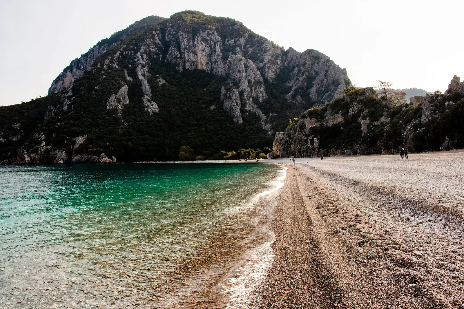 Het kiezelstrand van Çıralı met helder water en een beboste rotsheuvel